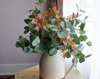 Könnte beinhalten: Eine cremefarbene Keramikkrug mit braunem Sockel enthält ein üppiges Arrangement aus grünen Eukalyptusblättern und roten Beeren. Der Krug steht auf einem hellen Beistelltisch aus Holz und schafft eine natürliche, einladende Ästhetik.