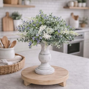 以下が含まれることがあります： 白い花と紫の花、緑の葉を使った装飾的なフラワーアレンジメント。 白いアンティーク調の台座と丸い木製トレイの上に置かれています。 背景は明るいキッチンです。