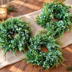 May include: Three circular wreaths constructed from green faux cedar foliage and accented with numerous small, light blue berries. The wreaths feature a dense, textured appearance with realistic needle-like branches and are arranged in a cluster.