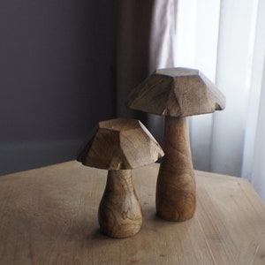 May include: Two wooden mushroom sculptures on a wooden table. The larger mushroom is brown and has a more rounded top. The smaller mushroom is also brown and has a more pointed top.