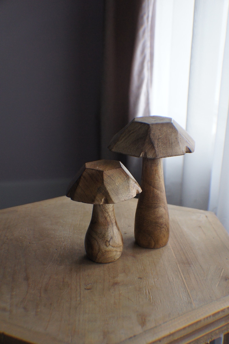 May include: Two wooden mushroom sculptures on a wooden table. The larger mushroom is brown and has a more rounded top. The smaller mushroom is also brown and has a more pointed top.