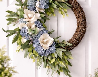 Corona de puerta de estilo rústico con magnolias e hortensias, adornada con follaje y hierbas.