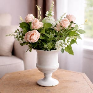 Könnte beinhalten: Ein Blumenarrangement mit pfirsichfarbenen Rosen, weißen Blüten und grünem Laub in einer weißen, urnenförmigen Vase. Die Vase steht auf einem sechseckigen Holztisch. Der Hintergrund umfasst ein Sofa und Vorhänge in neutralen Tönen.