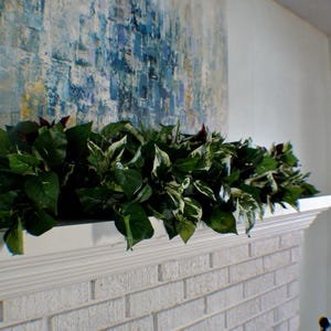 May include: A white brick fireplace with a white mantelpiece is decorated with a vibrant green garland. Above the mantelpiece hangs a large abstract painting in shades of blue and white, creating a calm atmosphere.