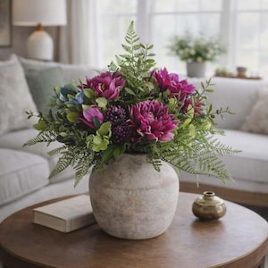 Puede incluir: Un arreglo floral en un jarrón texturizado de color claro. El ramo presenta dalias magenta vibrantes, tulipanes morados y follaje verde. El jarrón está sobre una mesa de centro redonda de madera oscura, con un pequeño recipiente decorativo de latón.
