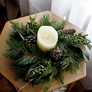 Puede incluir: Una vela de pilar color crema rodeada de un arreglo festivo de agujas de pino, piñas y bayas. La vela se encuentra sobre una mesa hexagonal de madera, creando un centro de mesa natural y de temporada.
