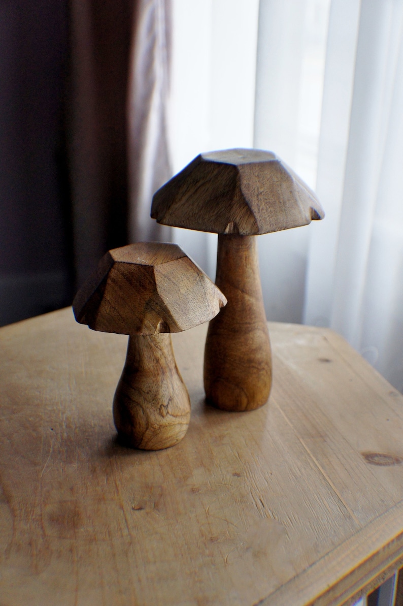 May include: Two wooden mushroom sculptures on a light brown wooden surface. The larger mushroom is brown and has a more rounded top. The smaller mushroom is a lighter brown and has a more angular top.