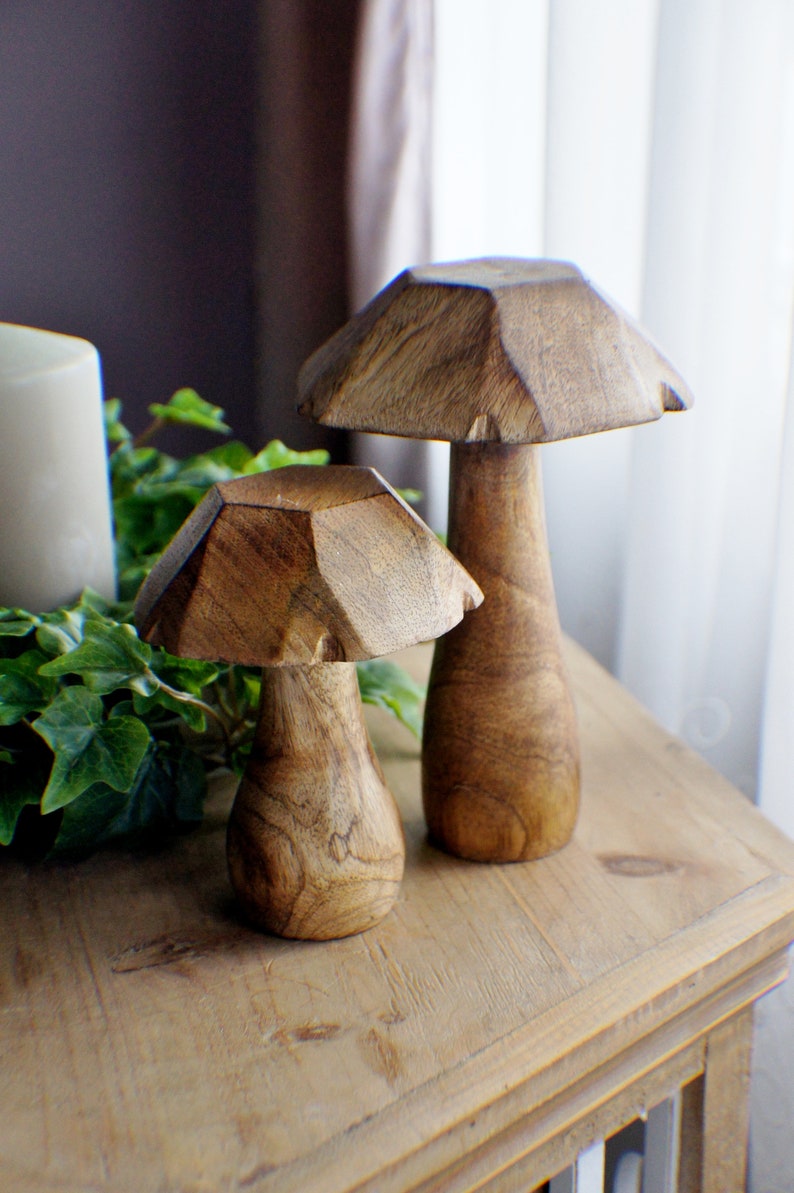 May include: Two wooden mushroom sculptures with a geometric design. The larger mushroom is brown and has a taller stem. The smaller mushroom is also brown and has a shorter stem. The mushrooms are sitting on a wooden table.