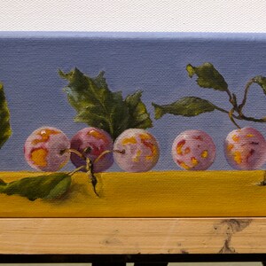 May include: A painting of a still life featuring six plums with pink and purple hues, arranged in a row on a yellow surface. The plums are surrounded by green leaves and branches against a light blue background.
