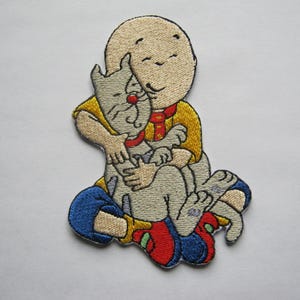 May include: Embroidered iron-on patch featuring a cartoon character with a bald head and a yellow shirt hugging a grey cat. The character has a red nose and a happy expression.