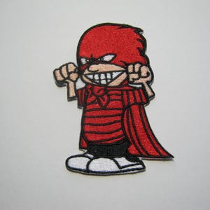 Peut inclure: Un écusson brodé représentant un personnage de dessin animé portant un masque rouge, une cape rouge et un t-shirt à rayures rouges et noires. Le personnage a un large sourire édenté, les poings serrés, un short noir et des chaussures blanches.