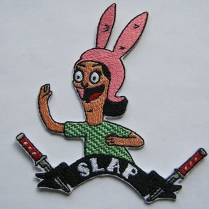 Pode incluir: Etiqueta bordada para passar a ferro com uma personagem de desenho animado com orelhas de coelho cor-de-rosa, uma camisa verde e a palavra "SLAP" em letras brancas sobre uma faixa preta com duas facas vermelhas e pretas.