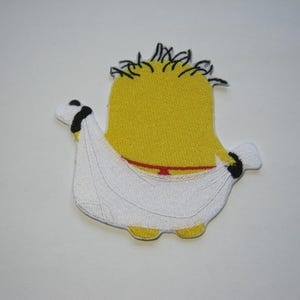 May include: Yellow felt minion character wearing a white felt toga. The minion has black felt hair and a red felt belt.