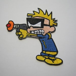 Peut inclure: Un écusson brodé représentant un personnage de dessin animé aux cheveux jaunes en pointe, portant des lunettes noires et un t-shirt bleu avec un éclair jaune. Le personnage tient un pistolet laser gris à bout orange, visant avec une expression intense.