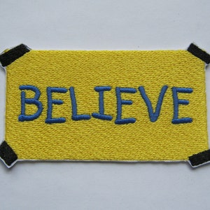 Peut inclure: Patch en tissu jaune avec du ruban adhésif noir autour des bords. Le patch a le mot "BELIEVE" brodé en fil bleu.