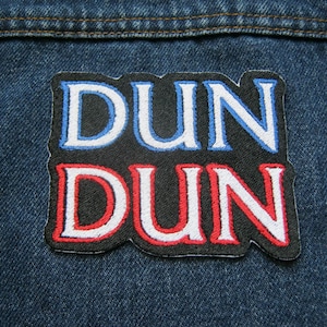 May include: A rectangular embroidered patch on dark blue denim fabric. The patch features a black background with the text DUN in light blue letters on the top line and the text DUN in red letters on the bottom line, both outlined in white.
