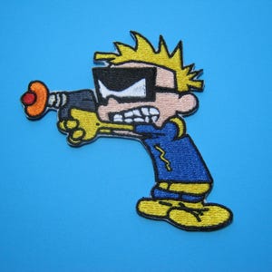 May include: An embroidered patch featuring a cartoon character with spiky yellow hair, wearing black goggles and a blue shirt, holding a dark gray ray gun with an orange tip. The character has a determined expression with clenched teeth.
