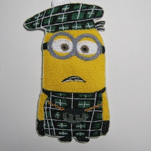Puede incluir: Un minion amarillo con un kilt de cuadros verdes y blancos y un gorro a juego. El minion tiene ojos grandes y redondos y una pequeña boca.
