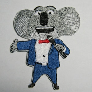 Puede incluir: Parche bordado para planchar con un koala que lleva un traje azul y una pajarita roja, sosteniendo un micrófono. El koala tiene una cabeza grande y gris con una amplia sonrisa.