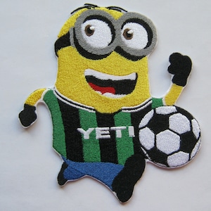 May include: A yellow minion cartoon character wearing a green and black striped soccer jersey with the word "YETI" on the front. The minion is holding a black and white soccer ball and is running.