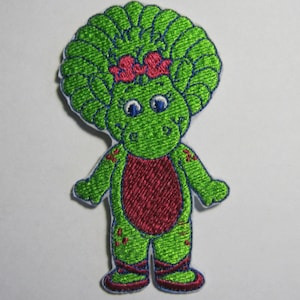 Pode incluir: Etiqueta bordada verde de um dinossauro de desenho animado com um laço rosa, vestindo um vestido rosa e sapatos verdes.