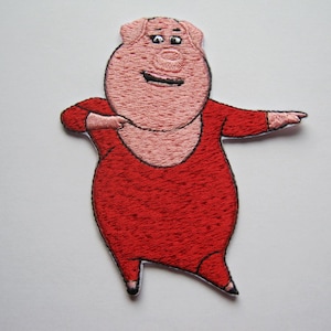 Pode incluir: Patch bordado com um porco de desenho animado em roupa vermelha, com os braços estendidos. O porco tem rosto e corpo rosa, com contorno preto. O patch foi projetado para ser passado a ferro ou costurado no tecido.