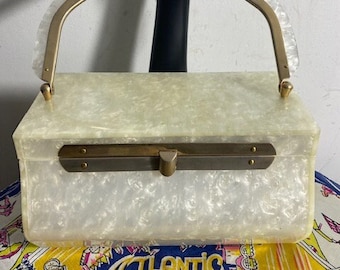 May include: A vintage rectangular handbag with a pearlescent finish. It has a gold-toned handle and a clasp closure. The bag is set against a background with a black hand sculpture and a colorful fabric with "Atlantic City" text.