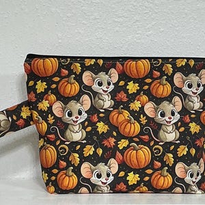 May include: A zippered pouch featuring a black background with a repeating pattern of cartoon mice, orange pumpkins, and autumn leaves in shades of yellow, orange, and red. The pouch includes a side wrist strap and a metal ring attachment.