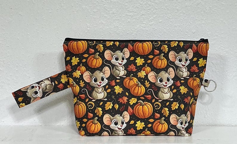 May include: A zippered pouch featuring a black background with a repeating pattern of cartoon mice, orange pumpkins, and autumn leaves in shades of yellow, orange, and red. The pouch includes a side wrist strap and a metal ring attachment.