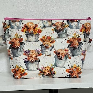 May include: A zippered pouch featuring a repeating pattern of Highland cows with floral crowns sitting in metal buckets. The fabric has a white background with colorful watercolor-style flowers and a bright pink zipper closure along the top edge.