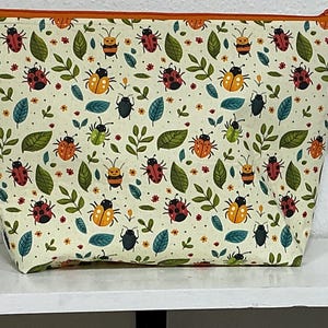 May include: A cream-colored zippered pouch with a colorful bug and leaf pattern. The design features various insects, green leaves, and small flowers. The zipper and trim are orange, and a silver ring is attached to the side.
