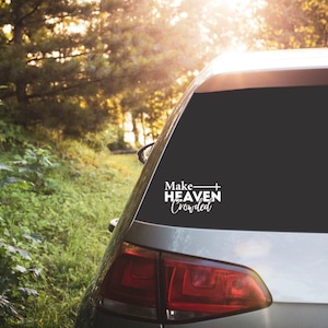 May include: A silver car with a black rear window. A white decal on the window reads "Make Heaven Crowded" in a script font. The car is parked on a dirt road with trees in the background.