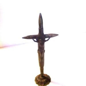 May include: A dark metallic crucifix sculpture constructed from bullet casings. The vertical post and horizontal crossbar are formed by ammunition cartridges, with a central figure attached to the front, all resting on a circular base.