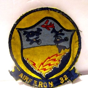 May include: Embroidered patch with a yellow border and a blue and yellow shield design. The shield features three blue airplanes, a red airplane, and a yellow and red lightning bolt design. The text "AIRE RON 32" is visible.