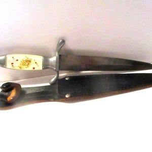 May include: A vintage dagger with a silver blade and handle. The handle is cream-colored with a gold design and red accents. The dagger is in a black leather sheath with a brown accent.