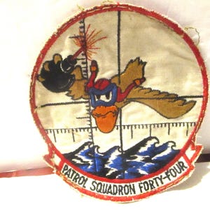 May include: Embroidered patch featuring a cartoon duck in a pilot's helmet and goggles, with a crosshair and the text "PATROL SQUADRON FORTY-FOUR". The patch has a red border and depicts waves and a laser beam.