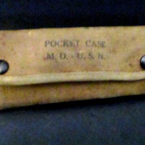 May include: A rectangular tan canvas pocket case with two dark metal snap buttons on the front flap. The text POCKET CASE M. D. - U. S. N. is printed in black ink on the center of the flap. The case features reinforced stitched edges.