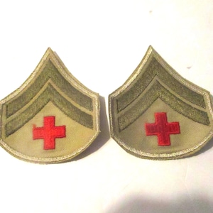 May include: Two matching embroidered patches featuring a tan background with two olive green chevrons pointing upward. Each patch displays a bright red cross symbol centered below the chevrons, finished with a tan stitched border edge.