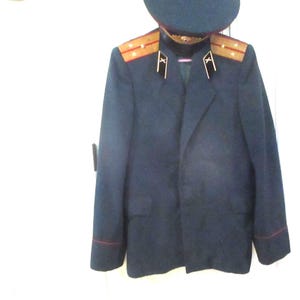 May include: A dark blue military-style jacket with a matching peaked cap. The jacket features shoulder boards with gold-colored stripes and stars, black collar tabs with white insignia, and thin red piping on the cuffs and collar.