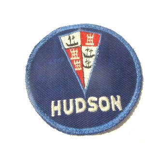 May include: A circular embroidered patch featuring a deep blue background with a light blue stitched border. In the center, a triangular shield is divided into red and white sections with black ship icons. The word HUDSON is stitched in white.