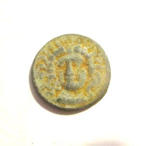 May include: A circular, weathered metal coin featuring a central embossed face with radiating lines. The surface displays a dull grey tone with patches of orange-brown oxidation and a textured, worn finish across the entire circular object.