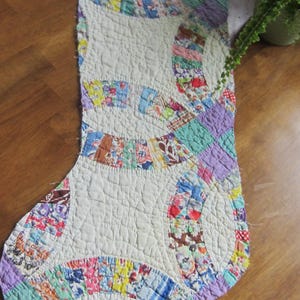 May include: A quilted Christmas stocking featuring a double wedding ring pattern. The design consists of off-white fabric sections interspersed with colorful, multi-patterned fabric strips in shades of blue, red, yellow, green, and purple.