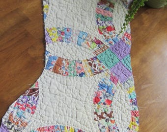 Op de afbeelding: Een gewatteerde kerstsok met een dubbel trouwringpatroon. Het ontwerp bestaat uit gebroken witte stoffen delen afgewisseld met kleurrijke, veelkleurige stoffen stroken in tinten blauw, rood, geel, groen en paars.