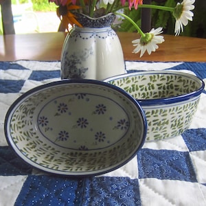 May include: Two ceramic bowls with blue rims and floral patterns. One is oval-shaped with blue flowers, the other is a round bowl with a green leaf design. A vase with flowers sits behind the bowls on a blue and white patterned tablecloth.