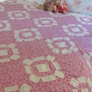 Puede incluir: Un edredón rosa y blanco con un estampado floral y formas geométricas blancas. Dos osos de peluche están sentados en el edredón, que está sobre una cama con almohadas con estampado floral.