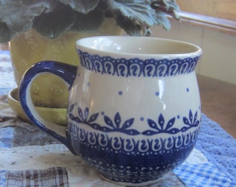 Op de afbeelding: Een keramische mok met een ronde vorm en een blauw handvat, voorzien van een crèmekleurige basis versierd met ingewikkelde donkerblauwe bloem- en geometrische patronen, inclusief een geschulpte rand bij de opening en een gestippelde band.