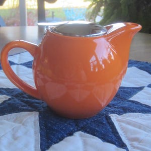 May include: A glossy orange ceramic teapot with a curved handle and a silver lid. The teapot sits on a blue and white quilted surface. The teapot is designed for brewing tea.