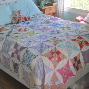 May include: A patchwork quilt featuring a repeating pattern of squares and curved shapes in various colors and prints, including red, blue, pink, yellow, and green, set against a cream-colored background, covering a bed with light blue pillows.