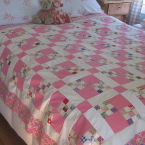 May include: A patchwork quilt featuring a repeating grid pattern of solid pink squares and white squares, with some squares containing multi-colored small-scale floral and geometric prints. The quilt has a pink and white patterned border.