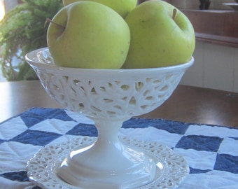 Puede incluir: Un cuenco blanco adornado con pie, lleno de tres manzanas verdes. El cuenco se encuentra sobre un plato a juego, sobre un edredón a cuadros azules y blancos. Las manzanas tienen tallos marrones.